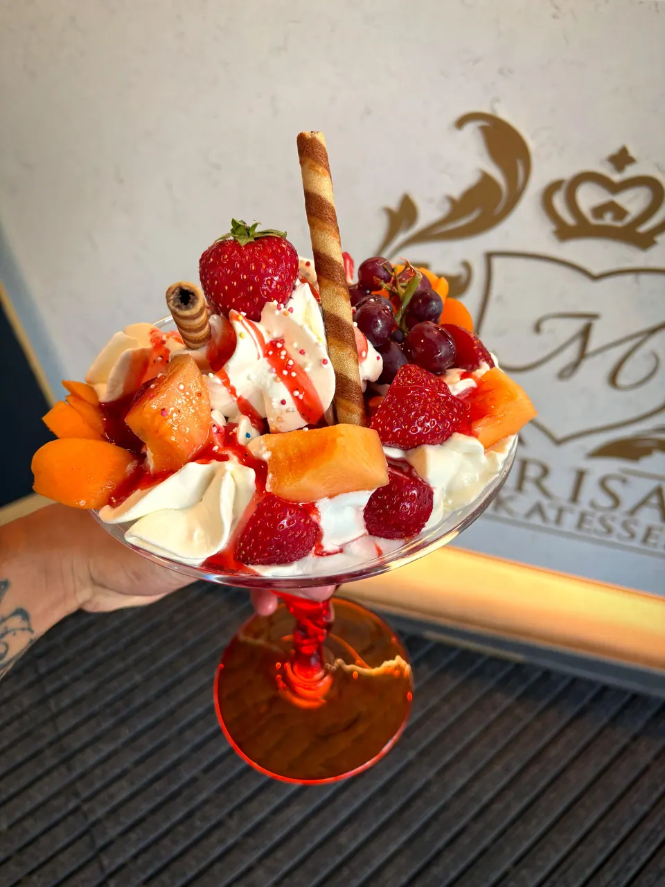 Eisbecher mit Erdbeeren, Waffelröllchen und Erdbeersoße vor dem Marisas-Wappen