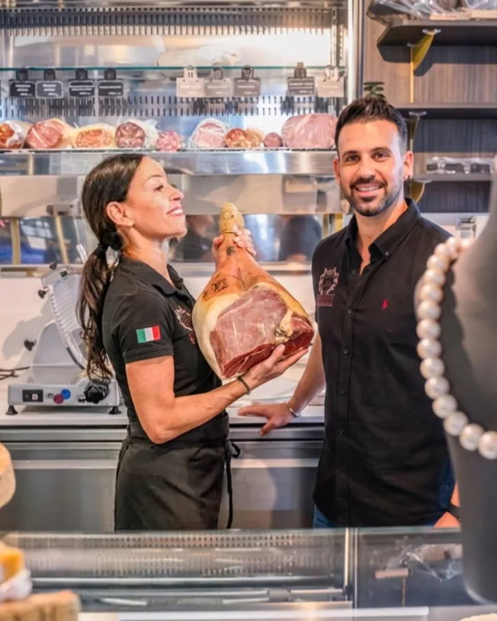 Marisa mit einem ganzen Prosciutto im Arm und Giorgio neben ihr, dahinter die Wurst- und Schinkenvitrine